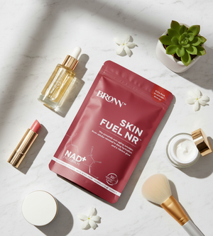 Cosmetic products including a red package labeled 'BRONN Skin Fuel NR', a lipstick, and a brush on a light surface with flowers and a plant.