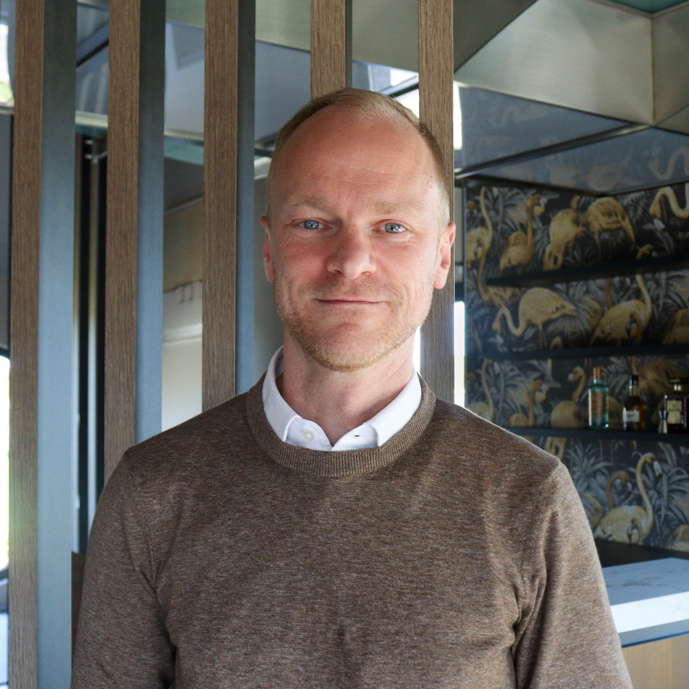 Man wearing a brown sweater and white shirt in an indoor setting with wooden panels and decorative wall art.