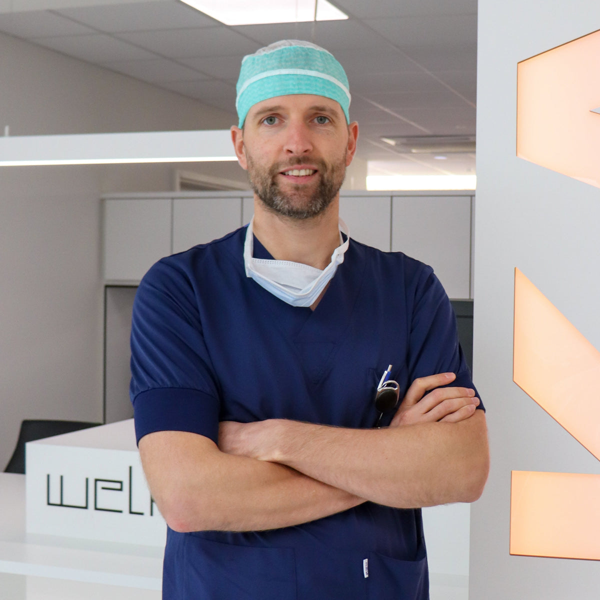 Doctor in blue scrubs with a teal scrub cap standing in a medical office.
