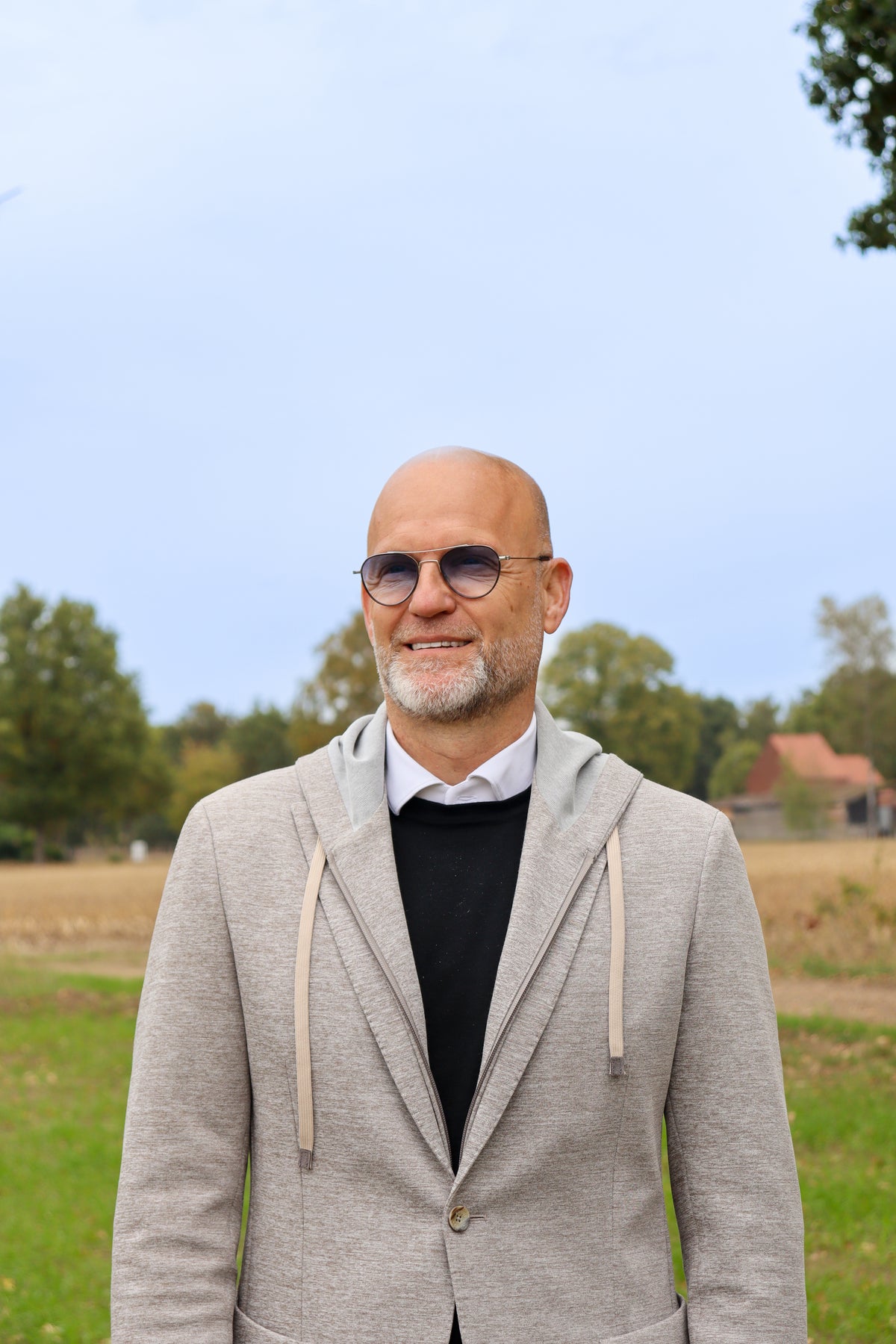 Man wearing sunglasses and a gray blazer standing in an outdoor setting with trees and a clear sky.