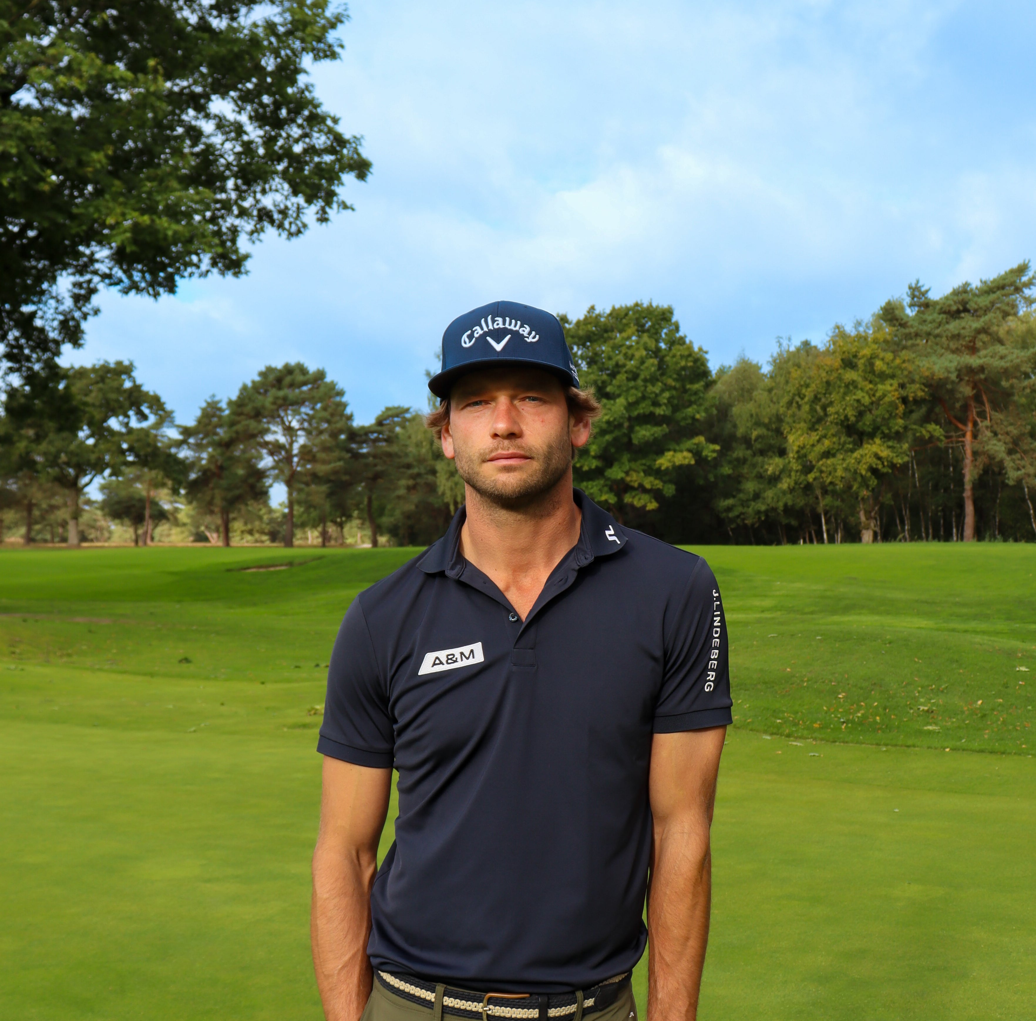 Man wearing a navy blue polo shirt and cap on a golf course with trees in the background