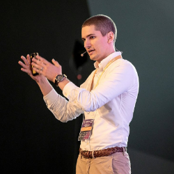 Man in a white shirt and beige jacket speaking on stage with a microphone.